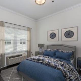 A modern bedroom with a bed covered in blue and patterned bedding, featuring matching pillows. Two pieces of artwork hang above the headboard. A lamp is situated on the bedside table near a window with blinds.