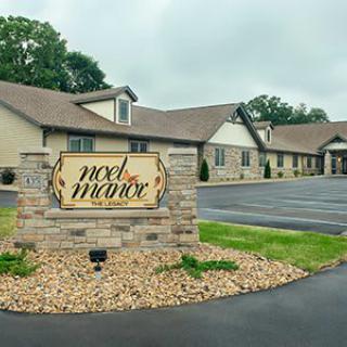 A view of a building with a sign reading 'Noel Manor' surrounded by greenery and a parking area.