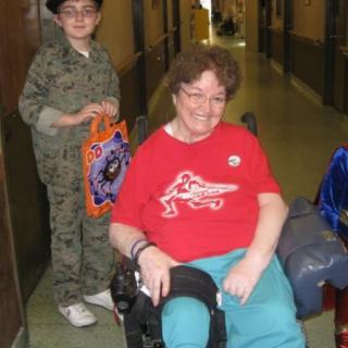 A child in a costume stands next to an elderly person in a wheelchair, both in a hallway.