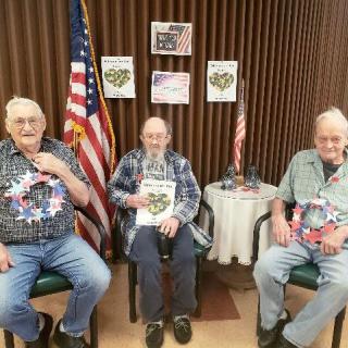 Three senior men are seated indoors, each wearing patriotic-themed attire with American flags in the background.