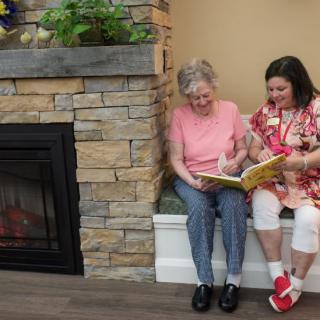 An elderly woman and a younger woman sit together by the fireplace, enjoying a book.