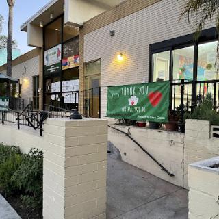 A view of a restaurant entrance featuring a green banner, outdoor seating, and potted plants.