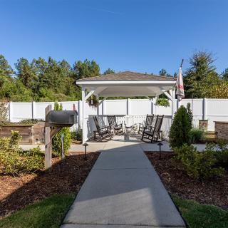 A cozy outdoor patio area with rocking chairs under a pavilion, surrounded by a landscaped garden and a clear blue sky.