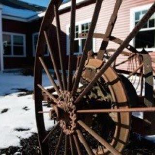 A rustic wagon wheel stands in the snow near a building.