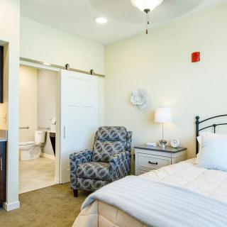 A cozy bedroom with white walls and a comfortable bed, featuring a patterned armchair, nightstand, and lamp.