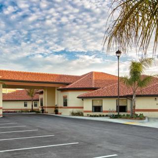 A spacious building with a red-tiled roof, surrounded by a parking lot and palm trees under a cloudy sky.