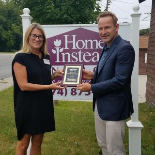 Two individuals exchanging an award in front of a Home Instead sign.