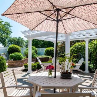 A sunny patio with a wooden table and chairs under a large umbrella, surrounded by lush green landscaping.