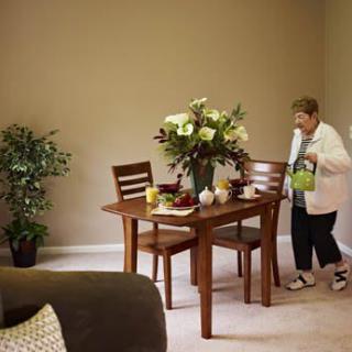 A dining area with a wooden table and chairs, adorned with a vase of flowers, while a person moves nearby.