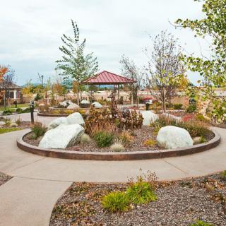 A scenic garden with a circular pathway surrounding a pavilion, featuring various plants and rocks.