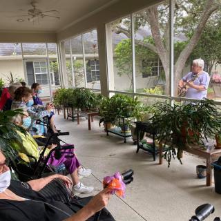 A musician performs outdoors for a group of elderly people sitting on a porch, maintaining social distance.