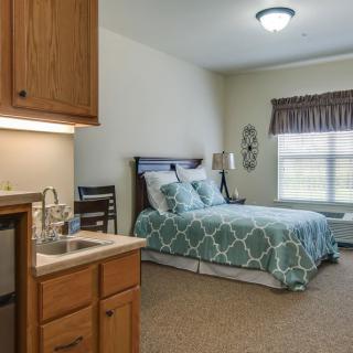 A cozy bedroom with a small kitchenette, featuring a neatly made bed and a window with curtains.