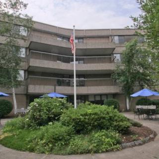 A multi-story apartment building with balconies, featuring a central flagpole and outdoor seating surrounded by landscaping.