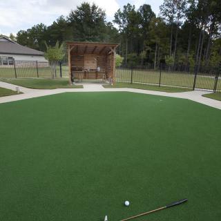 A well-maintained outdoor putting green with golf clubs and a ball, surrounded by fencing and a small shelter in the background.