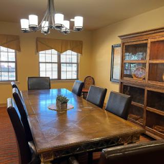 A spacious dining room featuring a large wooden table, black chairs, and a stylish chandelier, illuminated by natural light from wide windows.