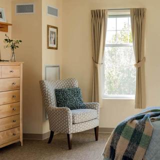A cozy bedroom featuring a patterned armchair with a blue cushion, a wooden dresser, and a window with beige curtains.