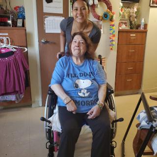 A caregiver smiles while standing behind an elderly woman in a wheelchair, showcasing a moment of companionship and care.