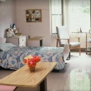 A tidy and bright hospital room with a single bed, a bedside table with flowers, and a window letting in natural light.