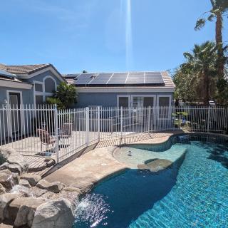A backyard features a swimming pool with a surrounding fence, solar panels on the house roof, and palm trees in the background.