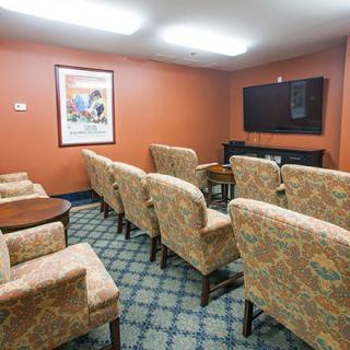 A cozy room with upholstered chairs facing a flat-screen television, adorned with a patterned carpet and a painting on the wall.