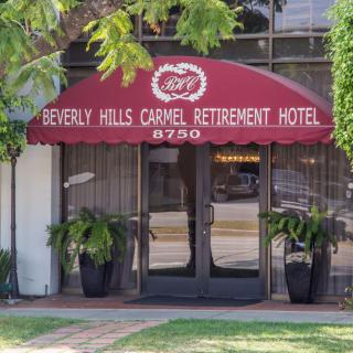Entrance to the Beverly Hills Carmel Retirement Hotel with a red awning and decorative plants.