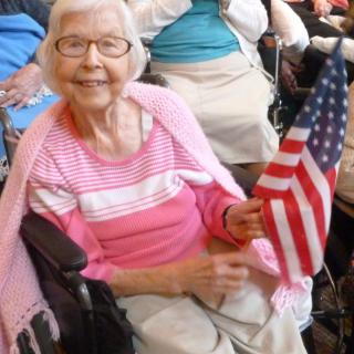An elderly woman in a pink sweater holds an American flag while seated in a wheelchair.
