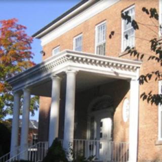 A historical brick building with a columned porch surrounded by trees.