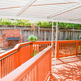 A sunny outdoor patio with a wooden deck, covered by a canopy and featuring a brick grill.