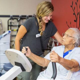 An elderly woman receives assistance from a caregiver during a rehabilitation session involving exercise equipment.