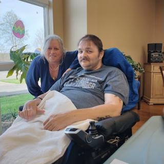 A patient in a wheelchair poses for a photo with a caregiver, both smiling in a bright, airy room.