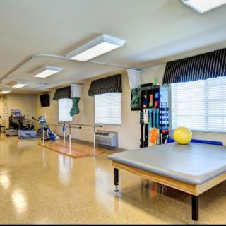 A spacious rehabilitation center featuring exercise equipment and a therapy table, well-lit with natural light from large windows.