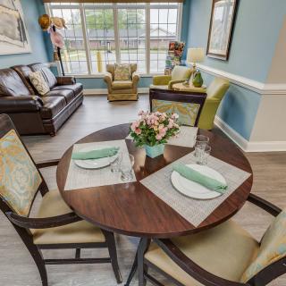 A cozy living room with a round dining table set for two, adorned with flowers, and comfortable seating near large windows.