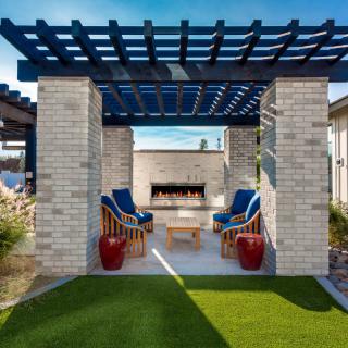 A modern outdoor patio with a pergola and fireplace, featuring cushioned chairs and blue accents.