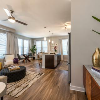 A spacious, modern living room with an open floor plan leading to a kitchen, featuring wood flooring and elegant decor.