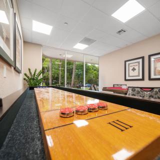 A cozy game room featuring a shuffleboard table, comfortable seating, and large windows overlooking greenery.