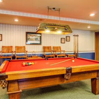 A well-lit game room featuring a classic pool table with red felt, surrounded by wooden chairs.