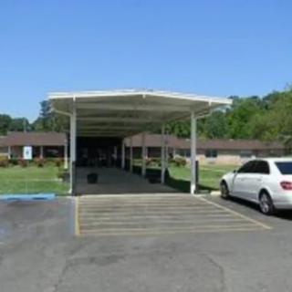 A car is parked under a carport in front of a building with surrounding greenery.