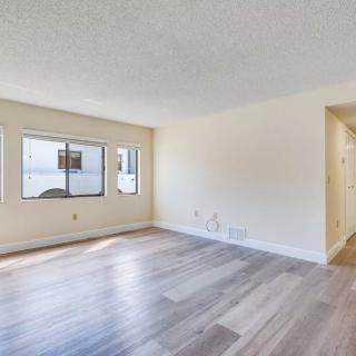 A spacious, empty living room with laminate flooring and a window leading to a hallway.