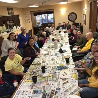 A large group of people gathered around a table covered with newspapers in a community setting.