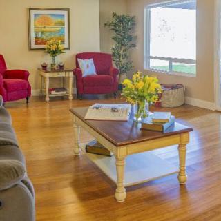 A cozy living room with wooden flooring, featuring red armchairs, a coffee table with flowers, and a large window letting in natural light.