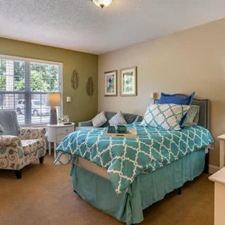 A cozy bedroom with a neatly made bed, decorated with blue and white bedding, a comfortable chair, and a large window letting in natural light.