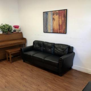 A cozy living room with a black leather sofa, a wooden piano, and colorful artwork on the wall.