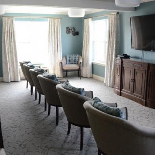 A cozy living room with rows of chairs facing a television, featuring large windows and elegant curtains.