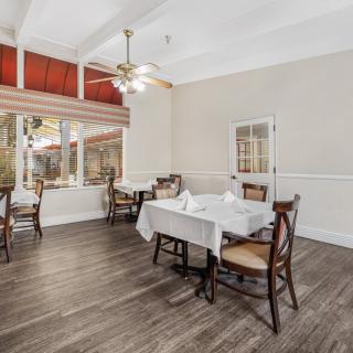 A well-lit dining room featuring wooden floors, tables with white tablecloths, and a ceiling fan.