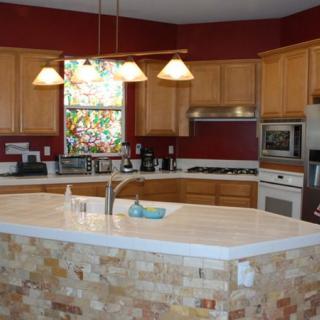 A modern kitchen with wooden cabinets, a large island countertop, and pendant lighting. A stained glass window adds a decorative touch.