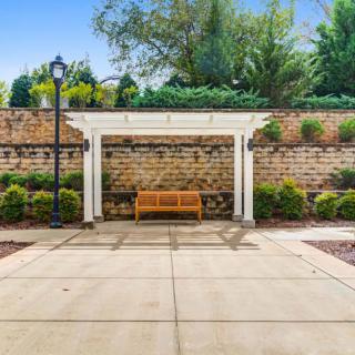 A wooden bench under a white pergola against a brick wall, surrounded by greenery and lit by a nearby lamp post.