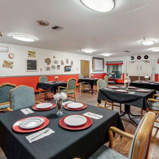 A cozy dining room with tables set for service, featuring black tablecloths and red-trimmed dinnerware.