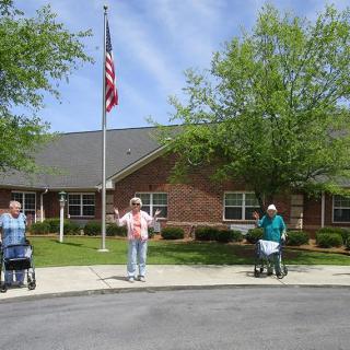 Three seniors with walking aids standing outside a building on a sunny day, with an American flag in the background.