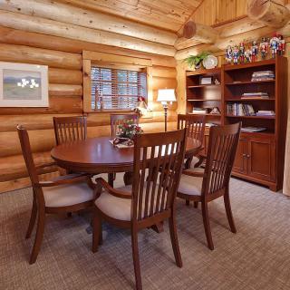 A cozy log cabin dining room with a wooden table and chairs, surrounded by warm wood-paneled walls and a well-stocked bookshelf.