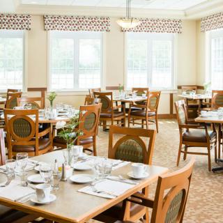 A well-lit dining room with neatly set tables and wooden chairs, featuring large windows that allow natural light to fill the space.